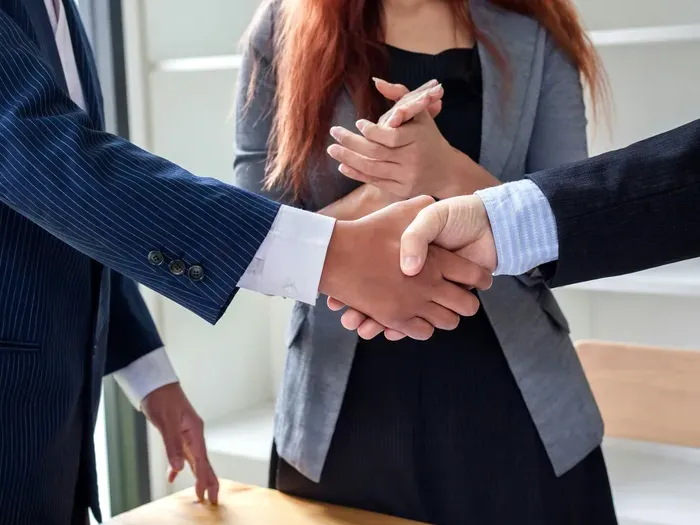 Businesspeople in suits shake hands as a colleague claps, confirming a partnership during an office meeting